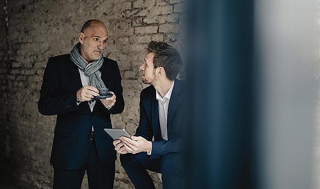 Zwei Männer im Anzug, einer sitzt mit einem Tablet in der Hand, der andere steht neben ihm mit einem Espresso und sie unterhalten sich.