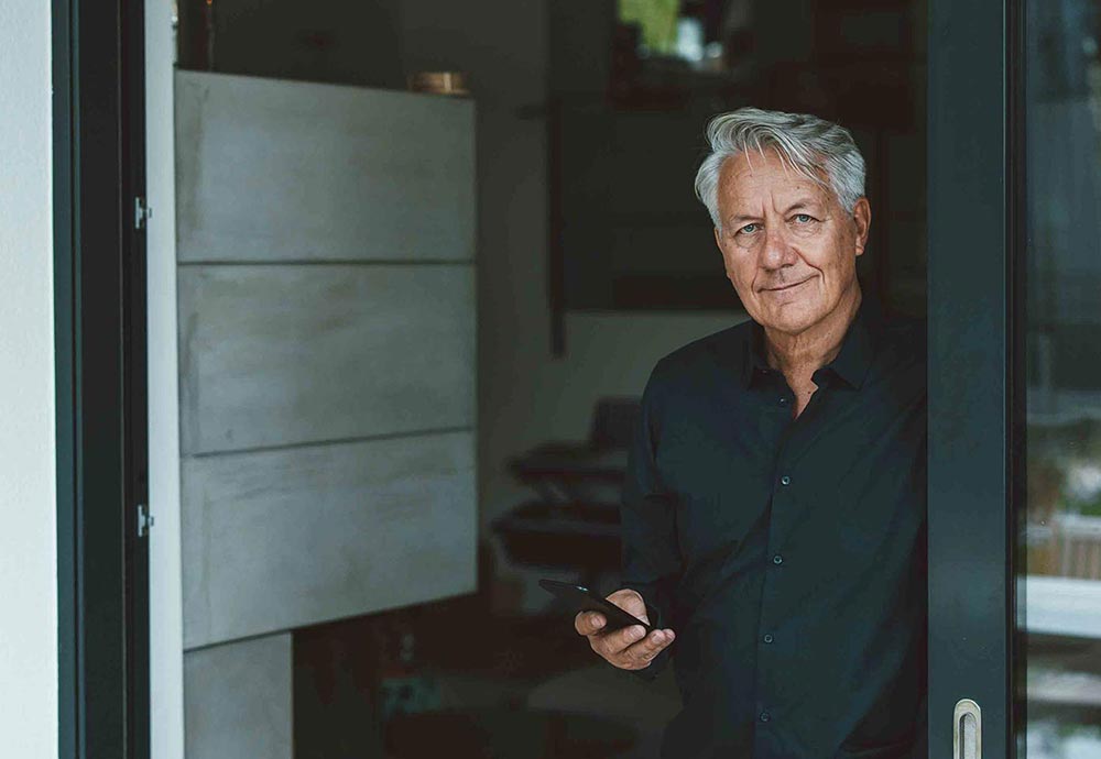 Ein Mann im Hemd steht im Glas-Türrahmen und hält sein Handy in der Hand