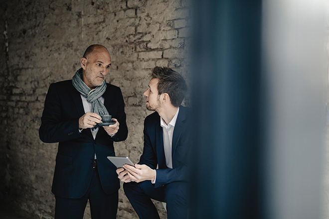 Zwei Männer im Anzug, einer sitzt mit einem Tablet in der Hand, der andere steht neben ihm mit einem Espresso und sie unterhalten sich.