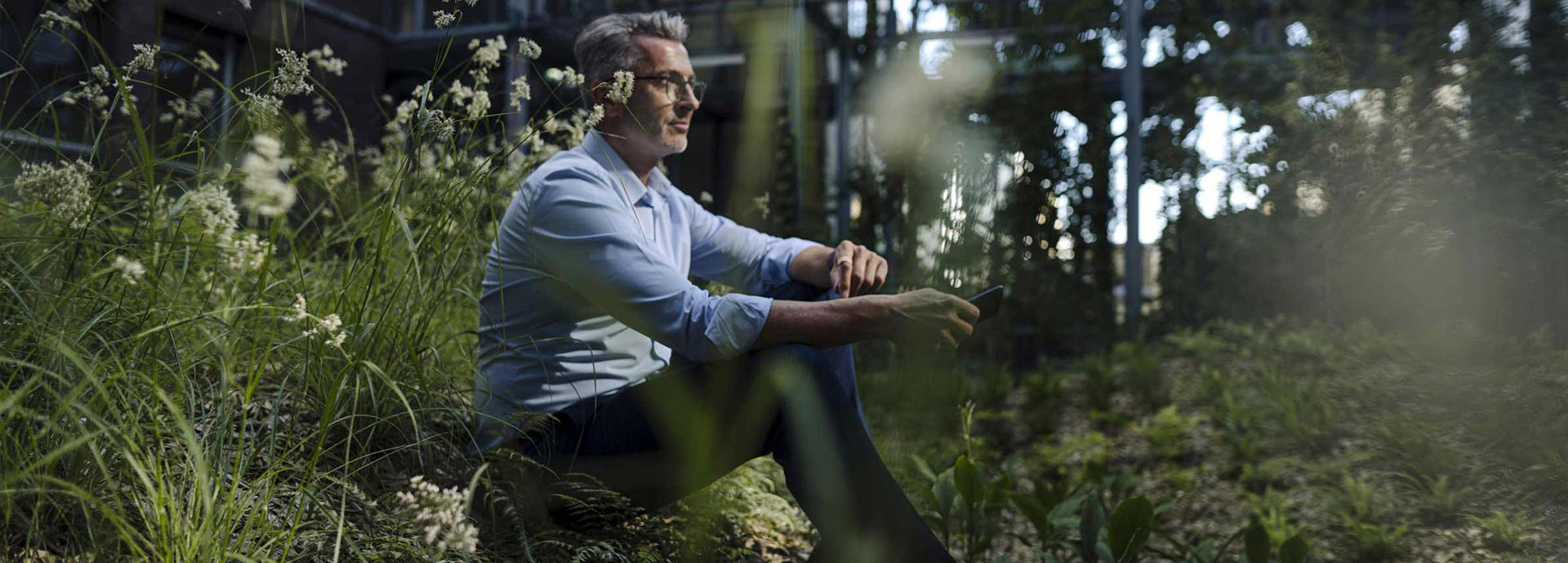 Ein Mann im Hemd sitzt auf der Wiese vor seinem Haus im Wald und hält sein Smartphone in der Hand.