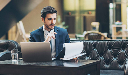 Ein Mann im Anzug sitzt auf einer Couch, mit einem Laptop vor sich. In seiner Hand hält er ein Notizbrett und blickt konzentriert darauf.