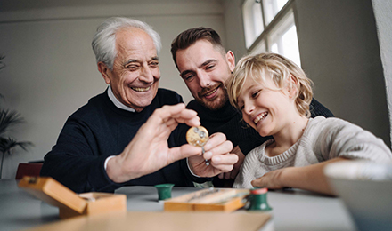 Drei Generationen, Großvater, Vater und Kind, lachen gemeinsam und schauen auf eine Münze, die der Großvater in der Hand hält.