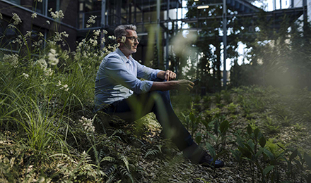 Ein Mann im Hemd sitzt auf der Wiese vor seinem Haus im Wald und hält sein Smartphone in der Hand.