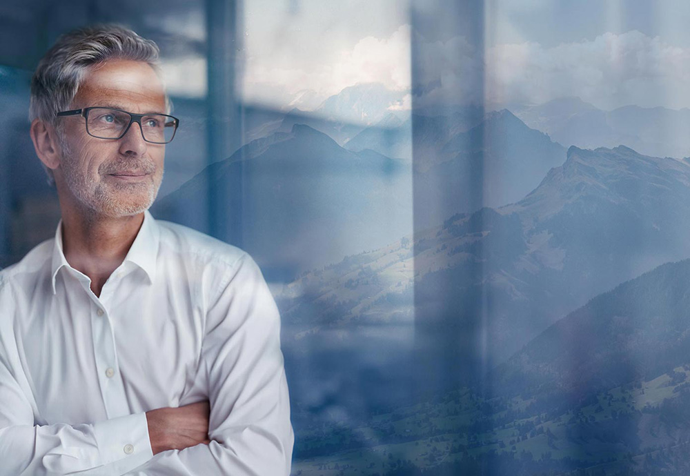 Ein Mann schaut mit einem leichten Schmunzeln durch eine Glasscheibe, im Hintergrund sind die Schweizer Berge zu sehen.