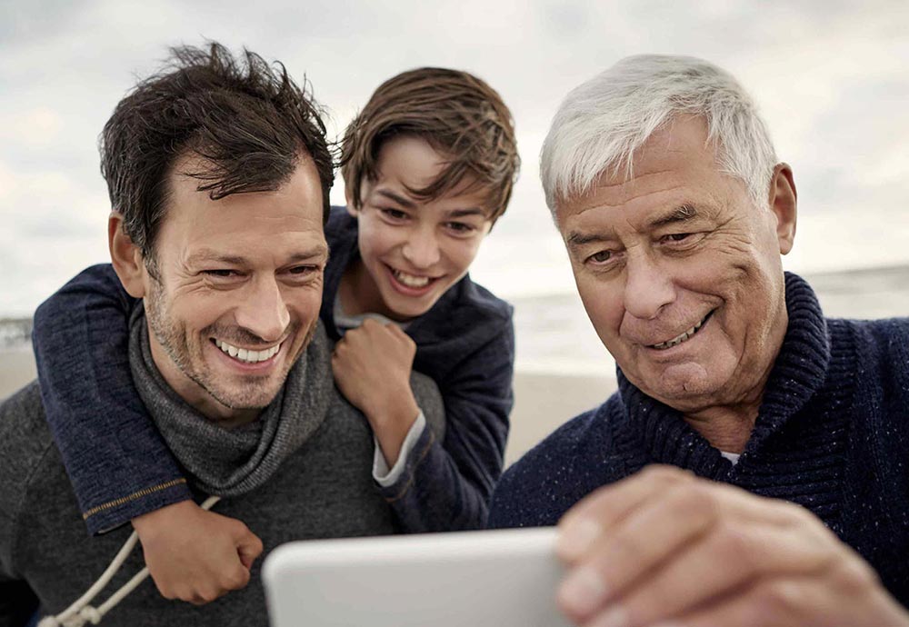 Ein Junge sitzt auf dem Rücken seines Vaters, neben ihm steht der Großvater. Gemeinsam schauen sie auf ein Handy und lachen.