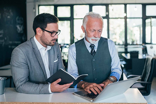 Ein Mann und ein älterer Mann sitzen nebeneinander an einem Schreibtisch. Der jüngere Mann hält ein Notizbuch in der Hand, und beide schauen gemeinsam in den Laptop.
