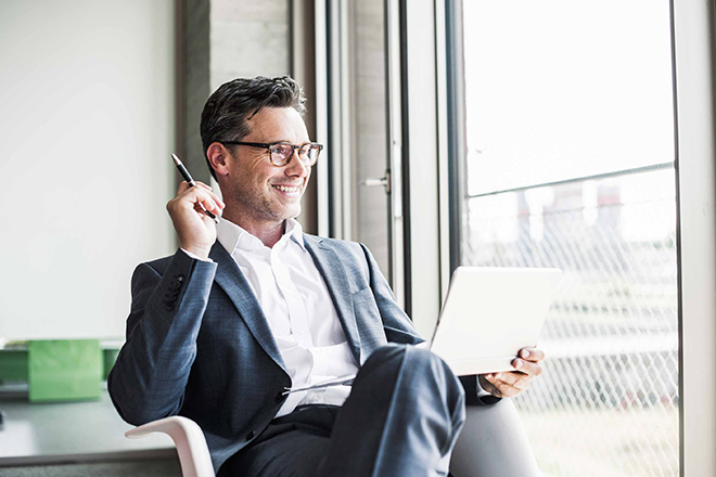 Ein Mann im Anzug, lächelnd und entspannt, sitzt auf einem Stuhl am Fenster, mit einem Bein überschlagen und einem Kugelschreiber sowie einem Laptop jeweils in einer Hand.