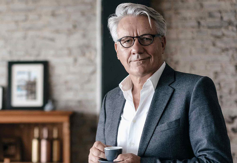 Ein Mann im grauen Anzug schaut schmunzelnd in die Kamera und hält eine Espresso-Tasse in der Hand.