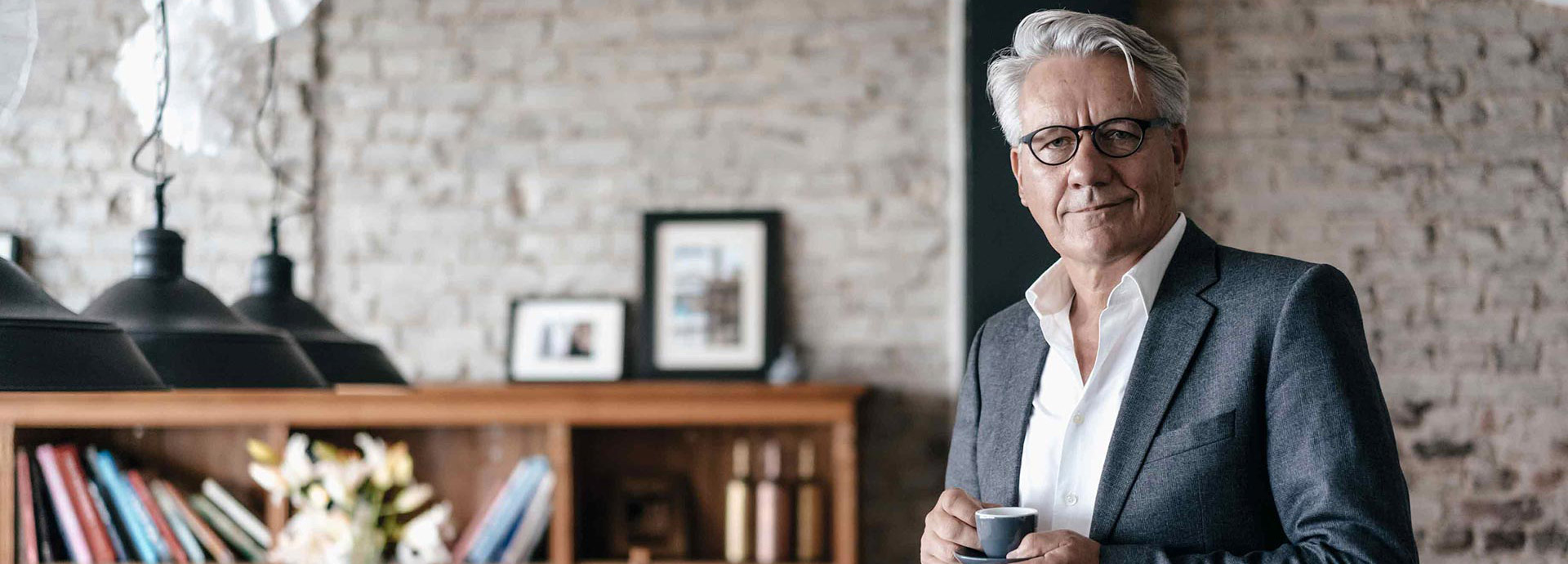 Ein Mann im grauen Anzug schaut schmunzelnd in die Kamera und hält eine Espresso-Tasse in der Hand.