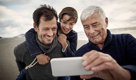 Ein Junge sitzt auf dem Rücken seines Vaters, neben ihm steht der Großvater. Gemeinsam schauen sie auf ein Handy und lachen.Ein Junge sitzt auf dem Rücken seines Vaters, neben ihm steht der Großvater. Gemeinsam schauen sie auf ein Handy und lachen.